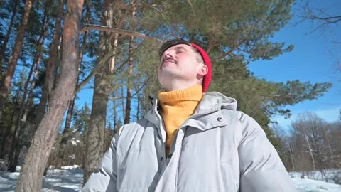 A man in a gray jacket and red hat stands in a snowy area surrounded by trees. Видео 337101770