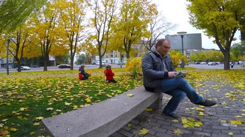 A man in a gray jacket sitting in the park on a granite bench and read e-books. Video stock 56342651