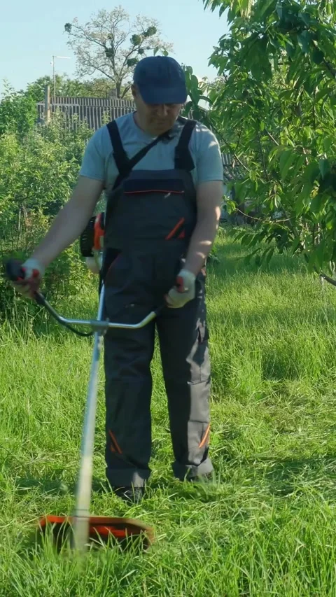 Man in gray overalls operates a string trimmer to cut grass in a lush garden 库存影片 338412230