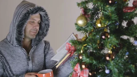 A man in a gray sweater drinks a hot drink. He enthusiastically reads the book Vídeo Stock 94853093