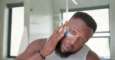 Man in gray tank looking down applying blue undereye hydrogel patches at vanity Stock Footage 336658739