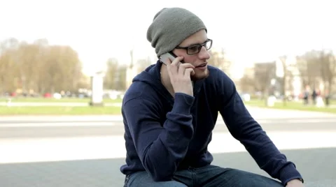 Man in grey hat and glasses sitting near the street, talking thru the phone. (1) Stock Footage 49535846