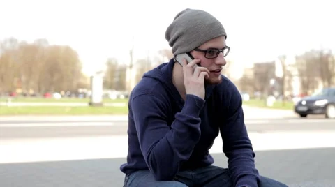 Man in grey hat and glasses sitting near the street, talking thru the phone. (1) Video stock 49535900