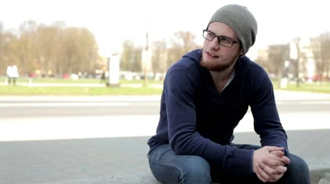 Man in grey hat and glasses sitting near the street and look around (3) Video stock 49535976