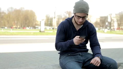Man in grey hat and glasses sitting near the street,writing messages on phone Stock Footage 49536092