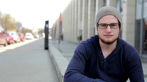 Man in grey hat and glasses sitting near building. He is looking into camera.. Stock Footage 49537222