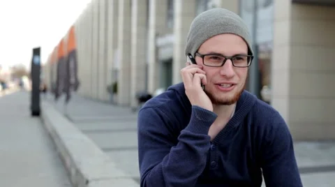 Man in grey hat and glasses sitting near building and talking thru the phone Video stock 49537236