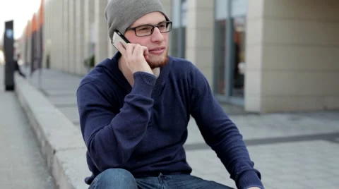 Man in grey hat and glasses sitting near building and talking thru the phone (40 Stock Footage 49537377