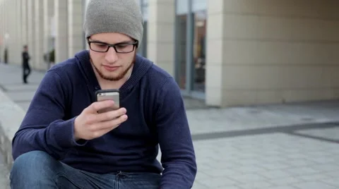 Man in grey hat and glasses sitting near building and writing text messages (43) Stock Footage 49537405