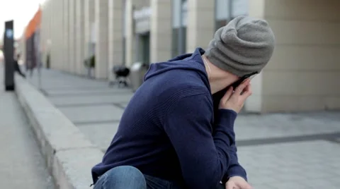 Man in grey hat and glasses sitting near building and talking thru the phone (40 Stock Footage 49537448