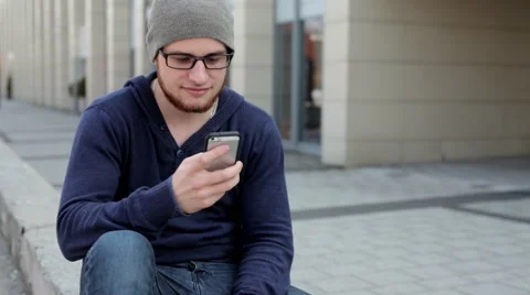 Man in grey hat and glasses sitting near building and writing text messages (43) Stock Footage 49537541