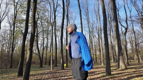 Man in a grey hat puts on a blue sports jacket in the forest during autumn Stock Footage 316833086