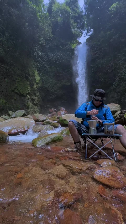Man Grinding Coffee Beans by Waterfall in Jungle River, Vertical Shot Stock Footage 321104778