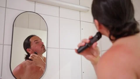 A man is grooming himself in front of a bathroom mirror while using an electric Stock Footage 317954894