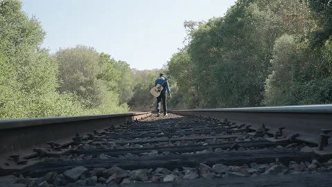 Man with guitar on back and bottle of whiskey in hand walking down train tra 스톡 동영상 235713615