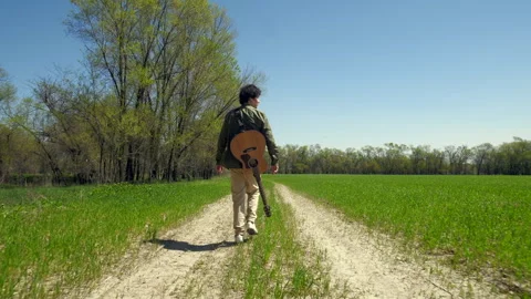 Man with a guitar walking down a rural road in summer along a narrow dirt road Vidéo 301402891