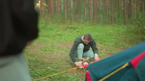 Man hammering peg into ground while person nearby jiggling another peg in forest Видео 310483386
