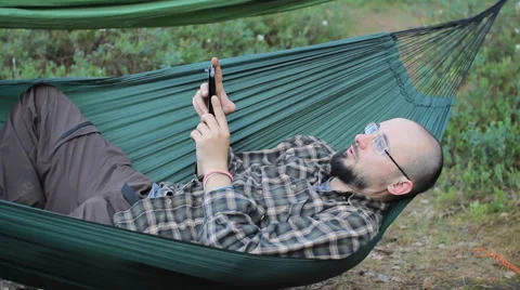 A man in a hammock is calling on the phone. Man in the woods with a beard and Stock Footage 64446465