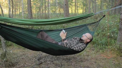 A man in a hammock is calling on the phone. Man in the woods with a beard and Stock Footage 64482017