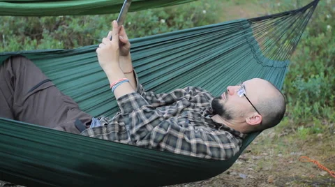 A man in a hammock making selfie on tablet. Man in the woods with a beard and Stock Footage 64449265