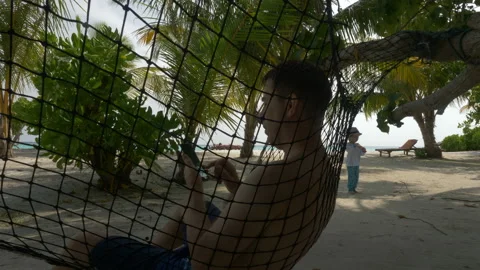 A man in a hammock with a mobile phone on the beach, in the background a girl Stock Footage 89998619