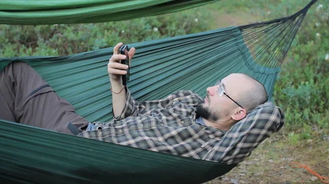 A man in a hammock touch phone. Man in the woods with a beard and glasses Stock Footage 64448511