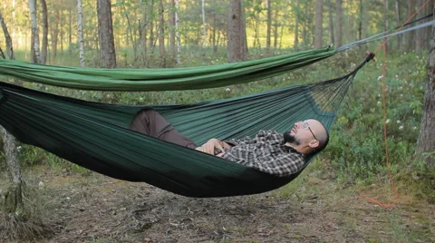 A man in a hammock touch smartwatch. Man in the woods with a beard and glasses Video stock 64484601