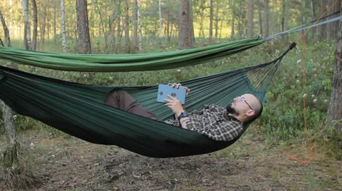 A man in a hammock touch tablet. Man in the woods with a beard and glasses Stock Footage 64484969