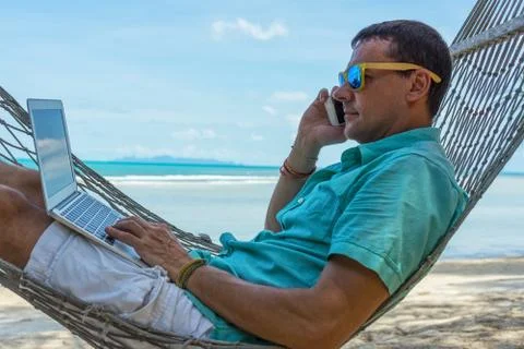 Man in a hammock on a tropical beach working on laptop and talking on the phone Stock Photos