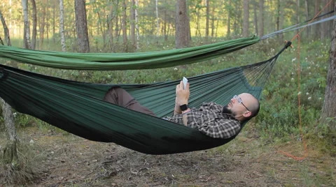 A man in a hammock is video calling on the phone. Man in the woods with a beard Stock Footage 64483424