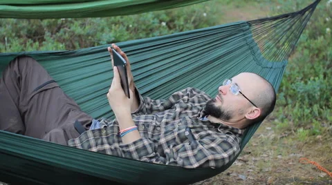 A man in a hammock is video calling on the tablet. Man in the woods with a beard Stock Footage 64450094