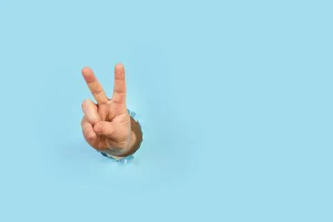 Man hand with victory gesture through a hole in a light blue background Stock Photos