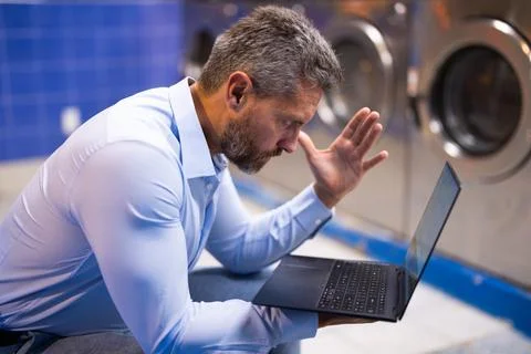Man handling housework and workload. Man doing laundry and working on laptop Stock Photos