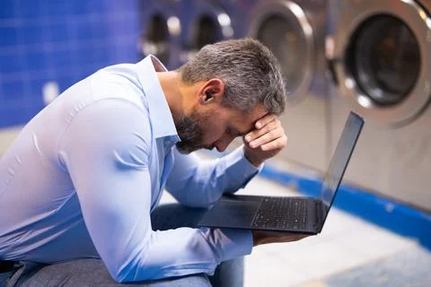 Man handling housework and workload. Man doing laundry and working on laptop Stock Photos