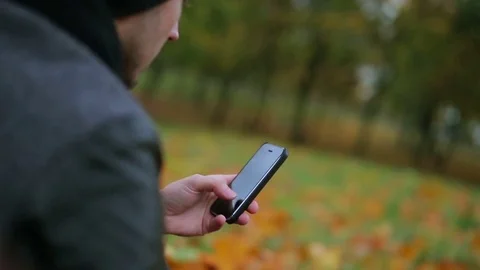Man hands using smartphone touchpad typing sms message in autumn park sitting on Stock Footage 71252526