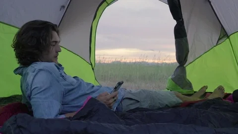 Man Hangs Out Inside His Tent And Checks His Smartphone, Sunset In Background Video stock 77053869