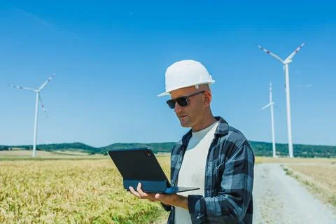 Man with hard hat uses tablet in a field with wind turbines on a sunny day Stock Photos