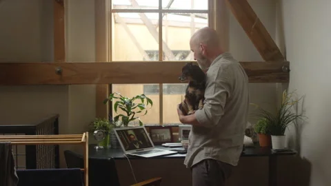 Man has a virtual happy hour while holding his cute dog while working from home 스톡 동영상 133295117