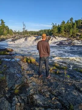 A man in hat admiring the view of a waterfall during sunset in the wilderness Stock Photos