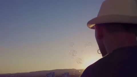Man in a hat, against the background of the sunset blowing bubbles, rear view Stock Footage 303570686