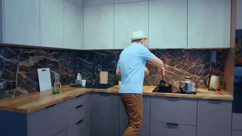 A man in a hat and a blue T-shirt, in a good mood, cooks and dances in the Stock Footage 181224967