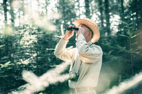 Man with hat and camera looking through binocular in forest. Stock Photos