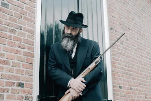 Man with hat and long beard standing with rifle against window shutters. Stock Photos
