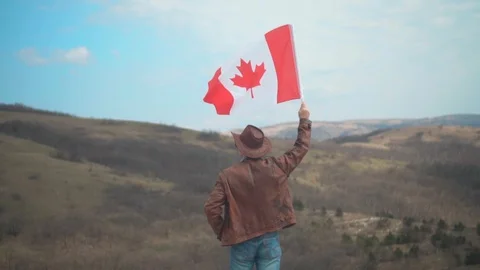 A man in a hat and sunglasses, leather jacket and jeans holding a Canadian flag. Stock Footage 107455641