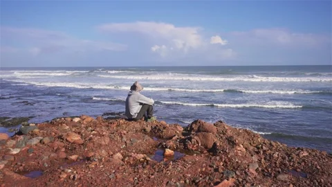 Man in hat and sunglasses sit and meditate on red rocky beach of ocean coast a Stock Footage 236386936