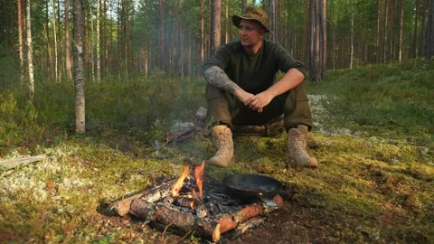 A man in a hat and tactical boots is sitting by the fire in the woods. Video stock 249618763