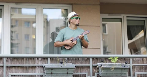 A man in a hat with a beard goes out onto the balcony and plays on the ukulele. Stock Footage 97226455
