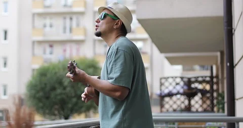 A man in a hat with a beard plays the ukulele on the balcony in the summer. Stock Footage 97379713
