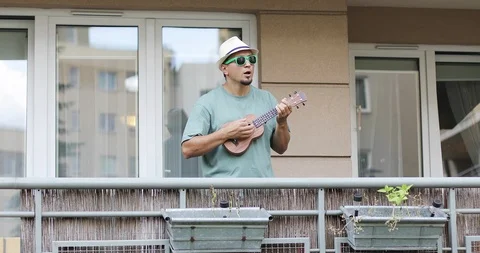 A man in a hat with a beard plays on the ukulele on the balcony. Stock Footage 98670151