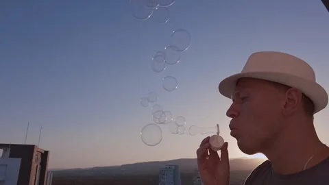 A man in a hat blowing soap bubbles, against the sky at sunset, camera tracking Stock Footage 309205397